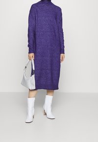 Lila stickad polokragnklänning, midilängd. Matchad med vita ankelstövlar och en silverhandväska. Texturen är mjuk och kraftig.