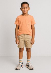 Kortärmad orange t-shirt med ficka, khakifärgade shorts, flerfärgade randiga strumpor och grå sneakers med blå och orange accenter.