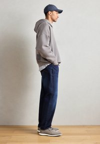 Grå hettegenser, blå denim jeans, grå sko. Uformell passform, myk tekstur, ingen synlige mønstre, står foran en nøytral bakgrunn.