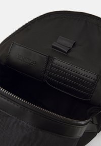 Černý nylonový vnitřek tašky se zipovým uzávěrem, obsahující kožený oddíl na karty a malou látkovou smyčku pro připevnění.