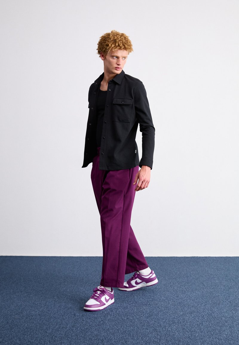 Camisa negra de manga larga con dos bolsillos en el pecho, combinada con pantalones sueltos morados y deportivas moradas y blancas, sobre una alfombra azul texturizada.