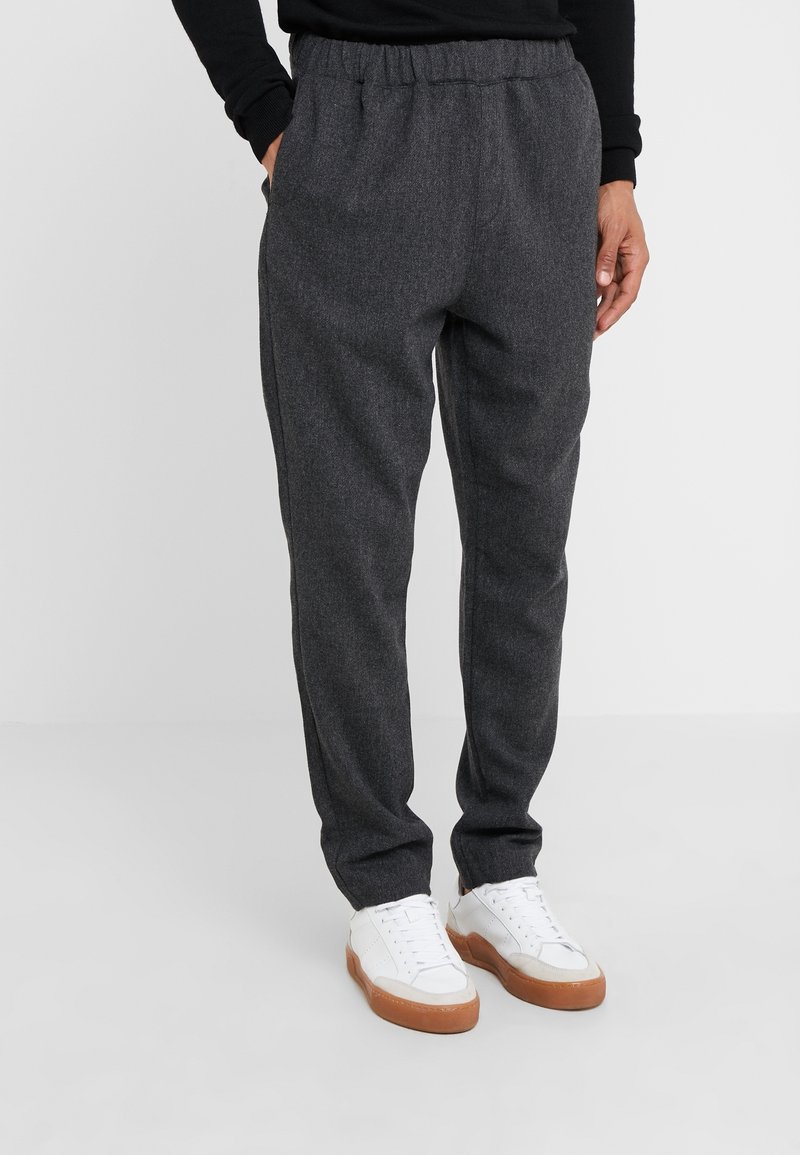 Homme portant un pantalon gris foncé et des baskets blanches avec des semelles marron, debout les mains dans les poches devant un fond uni.
