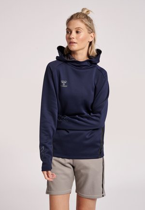 Kobieta z blond włosami upiętymi w koczek, ubrana w granatową bluzę z kapturem i jasnoszorty, stoi z jedną ręką skrzyżowaną, patrząc w prawo.