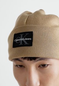 Beige gerippte Beanie-Mütze mit umgeschlagenem Saum, versehen mit einem schwarzen rechteckigen Patch, der das graue "Calvin Klein Jeans"-Logo zeigt.