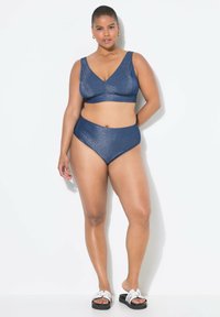 Bikini metallico blu navy con reggiseno a triangolo e slip a vita alta. Texture liscia con un leggero lucido. Indossato con sandali neri a ciabatta.