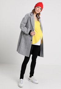 Casaco cinza em padrão houndstooth, blusa amarela em camadas, leggings pretas e ténis brancos. Gorro vermelho adiciona cor. O casaco tem um corte solto e fechamento com botões.