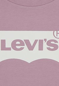 Levi's bijela oznaka izrezana na pozadini od ružičaste tkanine, prikazujući djelomični tekst "Levi's" i simbol registrirane robne marke.