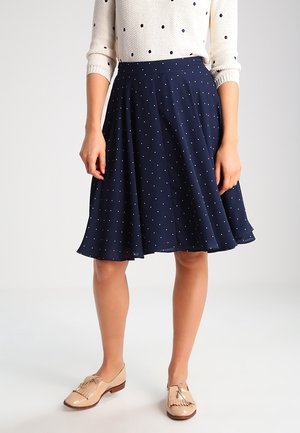 Jupe plissée marine avec un motif à pois blancs, en tissu léger. Associée à un pull crème et des chaussures beige.