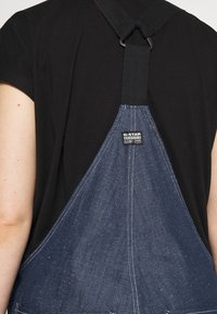 Jeansový overal tmavě modré barvy, viditelné švy, nastavitelný popruh a štítek G-Star. Nosený přes černé tričko.