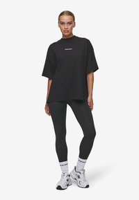 Tricou oversize negru cu guler rotund, text logo alb "PECADOR" pe piept, combinat cu colanți negri și adidași albi cu accente.