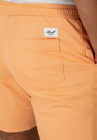 Shorts de color durazno hechos de algodón, con un único bolsillo trasero y una etiqueta de la marca. El tejido parece suave con una textura ligera.