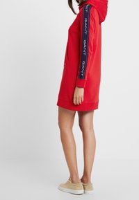 Rotes Kapuzenkleid aus weichem Stoff, mit einer Kängurutasche und blauen Ärmeln mit weißen "GANT"-Logopunkten.