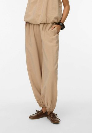 Luźne beżowe spodnie z elastycznymi mankietami noszone z brązowymi sznurowanymi butami i czarną bransoletką na lewym nadgarstku, ręce w kieszeniach.