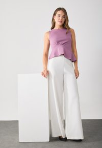 Woman leaning on a white rectangular pedestal wearing a sleeveless mauve peplum top, white wide-leg trousers, and black pointed shoes.