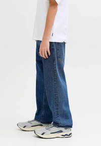 Kék, bő szárú farmernadrág, közepes mosással, látható varrással és két hátsó zsebbel, szürke és fekete sportcipővel párosítva.