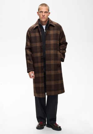 Homme portant un long manteau à carreaux marron, un pantalon noir, un pull noir zippé et des chaussures marron foncé avec des lacets rouges, debout avec une main dans la poche.