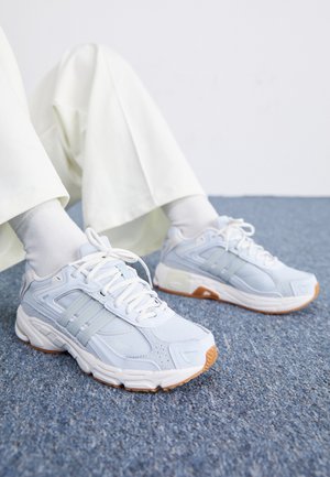 Persoon in witte broek en sokken, zittend op een blauwe tapijt, met lichte blauwe en witte sneakers met gumkleurige zolen en strikken die vastgebonden zijn.