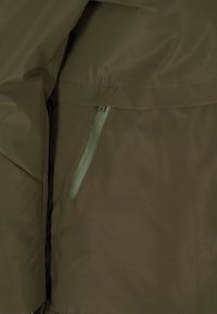 Kurtka w kolorze oliwkowej zieleni o eleganckim kroju, wykonana z fakturowanej tkaniny, z kontrastową kieszenią na zamek w jaśniejszym odcieniu zieleni.