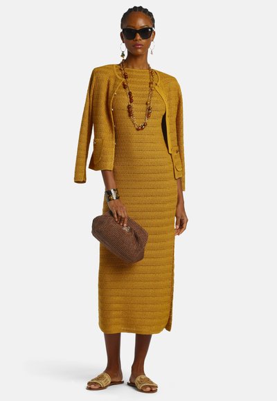 Frau trägt ein senfgelbes Strickkleid mit passendem Jackett, das über den Schultern drapiert ist, hält eine braune Clutch und ist mit einer groben Halskette sowie einer Sonnenbrille ausgestattet.