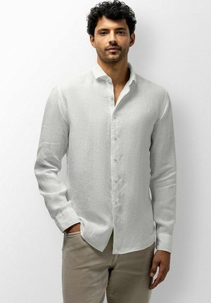 Man met donker krullend haar, gekleed in een witte blouse met knopen en lange mouwen en beige broek, staand tegen een effen witte achtergrond.