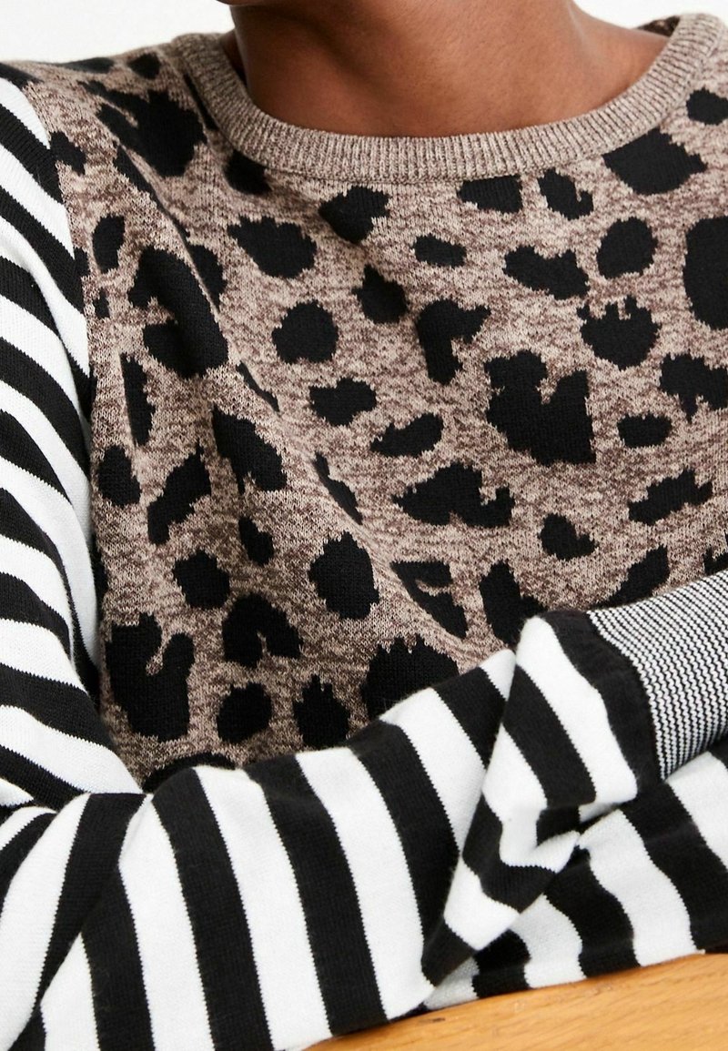 Personne portant un pull marron avec des taches léopard noires et des manches rayées noir et blanc, bras croisés sur une surface en bois.