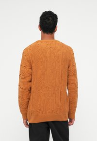 Υφασμάτινος πορτοκαλί πουλόβερ με έντονο σχέδιο πλεξίματος, στρογγυλή λαιμόκοψη και ριμπ τελείωμα στα μανίκια και το κάτω μέρος. Φοράει από πίσω.