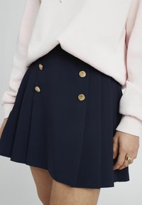 Mini-jupe navy en tissu texturé avec cinq boutons décoratifs dorés et un design plissé. Portée avec un sweat-shirt rose clair.