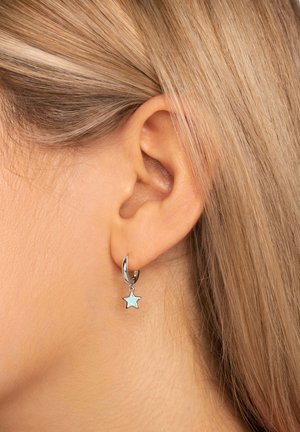 Silberner Creolenohrring mit einem kleinen, hellblauen Sternchen-Anhänger, der an der Unterseite hängt. Glatte Oberfläche, kompaktes Design, sichtbar am Ohr eines Models.