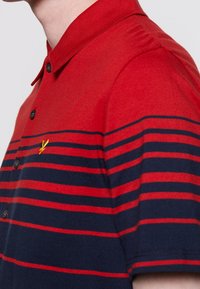 Polo shirt a righe rosse e navy con colletto, chiusura a bottoni e logo giallo ricamato sul petto. Tessuto dalla consistenza liscia.