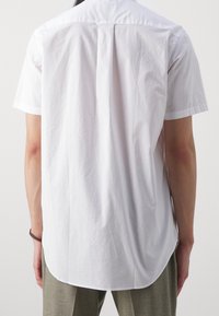 Personne portant une chemise blanche à manches courtes et un pantalon gris, debout, dos au spectateur, sur un fond uni.