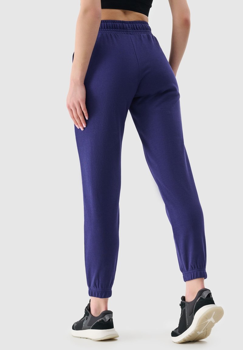Fioletowe dresowe spodnie z regulowanym paskiem w talii i elastycznymi mankietami, połączone z czarnymi butami sportowymi. Materiał wydaje się być miękki i elastyczny.