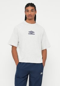 Mladenič z opletenimi lasmi nosi svetlo sivo majico Umbro in mornarsko modre hlače Umbro, roke ima v žepih in stoji pred belo ozadjem.