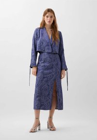 Lila bedrucktes Kleid mit einer kurzen Jacke, langen Ärmeln, vorderer Schlitz und Kordelzugdetails. Das Model trägt silberne Riemchensandalen.