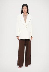 Biely dvojradový blejzer so širokými klopami, v kombinácii s béžovou košeľou s golierom a hnedými širokými nohavicami. Tmavé sandále s otvorenou špičkou.