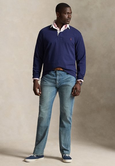 Man draagt een donkerblauw poloshirt met lange mouwen over een wit overhemd met kraag, lichtblauwe spijkerbroek, blauwe sneakers en een bruine riem, staand tegen een effen achtergrond.
