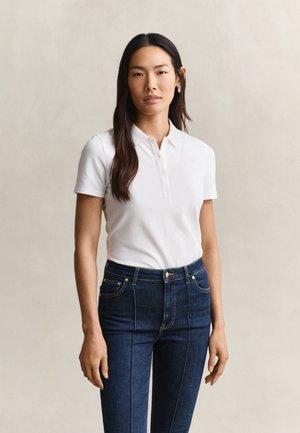 Femme aux longs cheveux foncés portant un polo blanc à manches courtes et un jean bleu foncé taille haute, debout devant un fond uni.