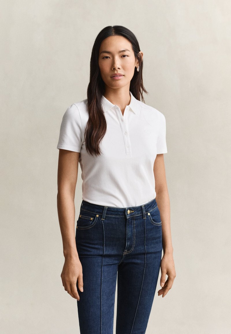Femme aux longs cheveux foncés portant un polo blanc à manches courtes et un jean bleu foncé taille haute, debout devant un fond uni.