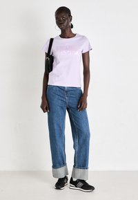 T-shirt corta viola con testo "pepe jeans"; abbinata a jeans in denim blu larghi e scarpe da ginnastica nere. Inclusa borsa a tracolla nera.
