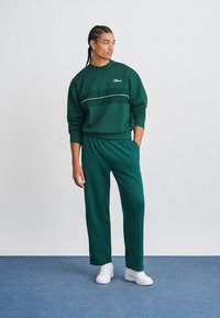 Πράσινη μπλούζα με λευκή λεπτομέρεια και λογότυπο, συνδυασμένη με ταιριαστά πράσινα παντελόνια φόρμας. Κατασκευασμένη από μαλακό υλικό, με χαλαρή εφαρμογή.