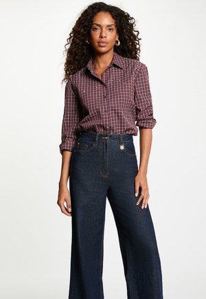 Mujer con cabello rizado que lleva una camisa de cuadros granate con mangas remangadas y vaqueros anchos de talle alto en azul oscuro.