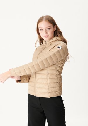 Donna che indossa una giacca trapuntata beige con cappuccio, che allunga il braccio in avanti su uno sfondo bianco uniforme.