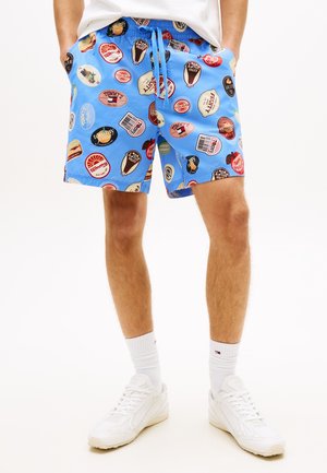 Man draagt blauwe korte broek met kleurrijke cirkelvormige fruitetiketpatronen, witte sokken, witte sneakers en een wit T-shirt, met handen in de zakken.