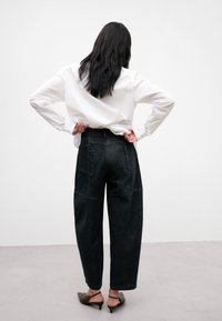 Tummat farkkukankaiset leveälahkeiset housut, joissa on korkea vyötärö, taka-taskut ja teksturoitu pinta, yhdistettynä valkoiseen nappikauluspaitaan.