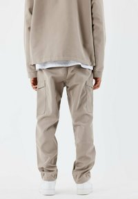 Pantalones cargo beige con puños elásticos y bolsillos laterales, combinados con una chaqueta beige holgada, llevada sobre una camisa blanca y zapatos blancos.