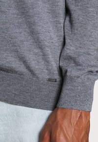 Gray knitted sweater with ribbed cuffs and hem. Features a small silver logo tag near the bottom hem. Soft texture, fitted design.