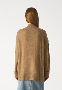 Neulepaita vaaleanruskeassa värissä, jossa on ribattu helma, korkea kaulus ja teksturoidut hihat. Ominaisuuksina leveä, rento istuvuus ja lasketut olkasaumat.