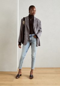 Blazer in suede grigio sopra un dolcevita nero, jeans in denim fitted azzurri, e sandali neri con tacco a blocco. Accessori minimalisti completano il look.