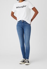 Camiseta blanca de manga corta con logo negro de "Wrangler", combinada con jeans ajustados azules y zapatillas negras con suelas blancas.