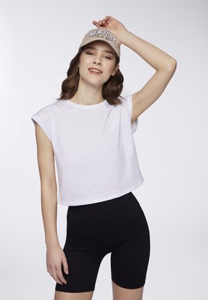 T-shirt bianca corta con maniche arrotolate abbinata a pantaloni ciclistici neri a vita alta. Il modello indossa un cappellino beige con la scritta "NEW YORK".