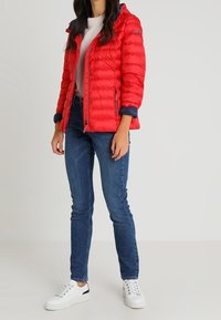 Vrouw in een rode pufferjack met een lichte trui, blauwe jeans en witte sneakers, staand tegen een neutrale achtergrond.